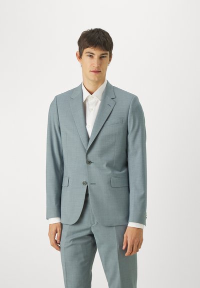 Costum bleu deschis din material texturat, cu rever în formă de notă, două butoane și un singur buzunar pe piept. Asortat cu o cămașă albă cu guler.