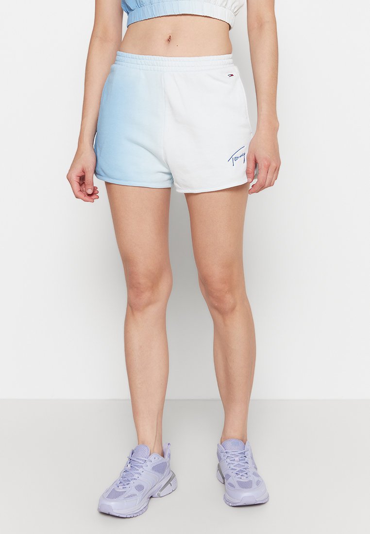 Pantaloni corti a gradiente bianco e azzurro chiaro con una cintura elastica e dettaglio del logo blu sul davanti, abbinati a scarpe da ginnastica lavanda.