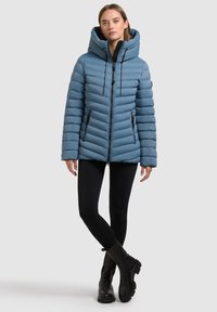 Giacca a vento azzurro chiaro con cappuccio, caratterizzata da una texture trapuntata, due tasche laterali con zip e dettagli in metallo nero.