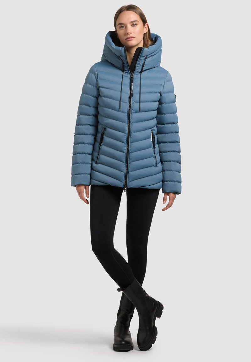 Giacca a vento azzurro chiaro con cappuccio, caratterizzata da una texture trapuntata, due tasche laterali con zip e dettagli in metallo nero.