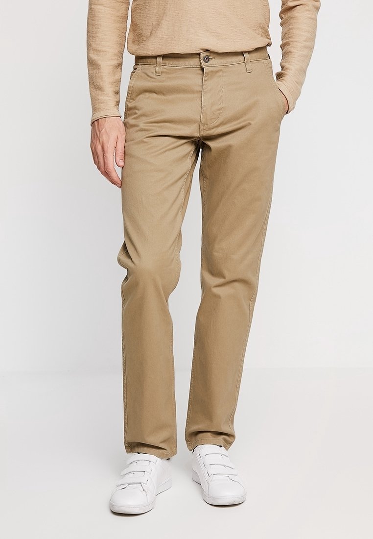 Uomo con pantaloni beige, camicia a maniche lunghe color sabbia e sneaker bianche con velcro, in piedi con una mano in tasca su uno sfondo bianco.