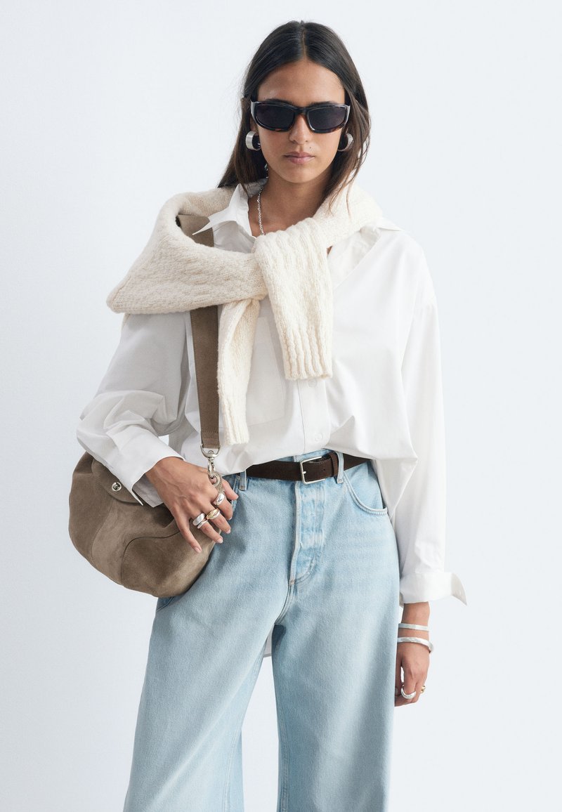 Femme portant des lunettes de soleil foncées, une chemise blanche, un jean bleu clair, un pull crème drapé sur les épaules et tenant un sac beige à l'épaule.