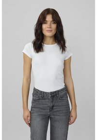 Žena so stredne dlhými tmavými vlasmi, oblečená v priliehavom bielom tričku s krátkym rukávom a sivých džínsoch s vysokým pásom, stojaca na jednoliatom sivom pozadí.