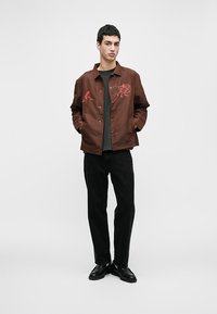 Jaqueta de lona castanha com desenhos em vermelho bordados no peito, usada sobre uma camisa preta e calças pretas, combinando com sapatos pretos.