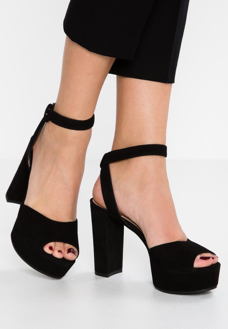 Escarpins à plateforme en daim noir à bout ouvert avec des brides à la cheville, portés avec un pantalon noir raccourci sur un sol clair.