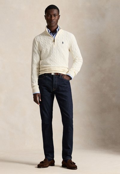 Uomo che indossa un maglione a trecce color crema sopra una camicia blu con jeans scuri e mocassini marroni con nappine, in piedi su uno sfondo semplice.