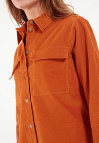 Chemise en velours côtelé de couleur rouille, avec un col boutonné, deux poches poitrine et une texture à côtes prononcée. Fermetures à boutons sur le devant.