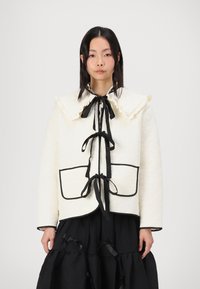 Λευκό καπιτονέ μπουφάν με μεγάλο ραμμένο κολάρο, μαύρες λεπτομέρειες και ζώνη στη μέση. Διαθέτει δύο μπροστινές τσέπες και λεία υφή.