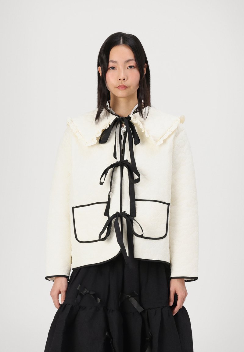 Λευκό καπιτονέ μπουφάν με μεγάλο ραμμένο κολάρο, μαύρες λεπτομέρειες και ζώνη στη μέση. Διαθέτει δύο μπροστινές τσέπες και λεία υφή.