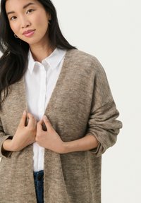 Beige neulecardigan, jossa on rento istuvuus, avoin etuosa ja pitkät hihat. Yhdistetty valkoiseen kaulukselliseen paitaan, jossa on näkyvää teksturoitua kangasta.
