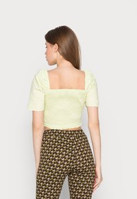 Top cropped verde claro com mangas bufantes e textura franzida; apresenta um decote quadrado nas costas. Combinado com calças castanhas com padrão.