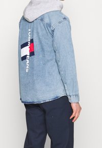 Jasnoniebieska koszula dżinsowa z szarym kapturem pod spodem. Posiada czerwony, biały i granatowy detal z logo na plecach oraz biały napis wzdłuż rękawa.