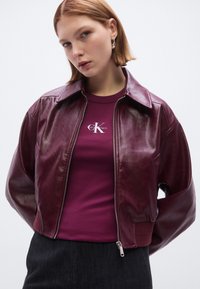 Bordeauxroter Leder-Bomberjacke mit durchgehendem Reißverschluss, weitem Kragen und gerippten Bündchen. Kombiniert mit einem eng anliegenden lila T-Shirt mit Logodruck.