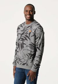 Grijze sweatshirt met een zwart bladenpatroon, ronde halslijn en geribde manchetten. Bevat een klein oranje logo op de borst. Gekoombineerd met een blauwe jeans.