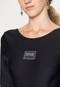 Svart långärmad topp i slät tyg med en rektangulär logotyp med texten "VERSACE JEANS COUTURE" och rund halsringning.