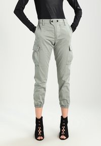 Lichtgrijze cargobroek met elastische boorden en twee zijzakken. Gestyled met een getailleerde zwarte top en zwarte veterschoenen met hak.