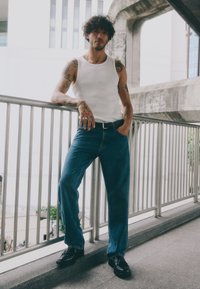 Valge tank-top, sinised denim teksad ja mustad kingad. Mees lokkis juustega seisab lõdvestunult piirdele toetudes kaasaegses linnakeskkonnas.