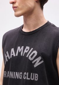 Νέος άντρας που φοράει μαύρο αμάνικο μπλουζάκι με ξεθωριασμένο κείμενο "CHAMPION TRAINING CLUB" εμφανές μπροστά.