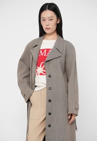 Μακρύ, ανοιχτό γκρι παλτό με γιακά με σχισμή και μαύρα κουμπιά, φορεμένο πάνω από λευκό πουκάμισο με κόκκινη γραφική εκτύπωση και μπεζ παντελόνι. Λεία υφή.