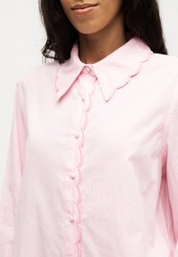 Femme portant un chemisier rose clair avec un col festonné et une bordure festonnée assortie le long de la ligne de boutons.