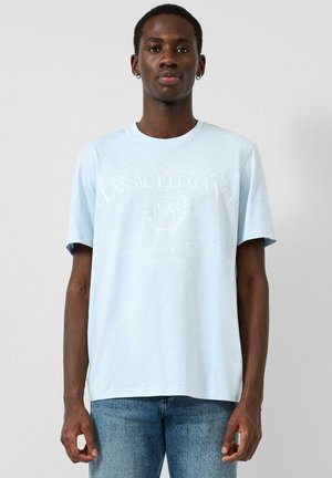 Młody mężczyzna w jasnoniebieskiej koszulce z nadrukiem i niebieskich jeansach, stojący na tle gładkiego jasnoszarego tła, zwrócony przodem.