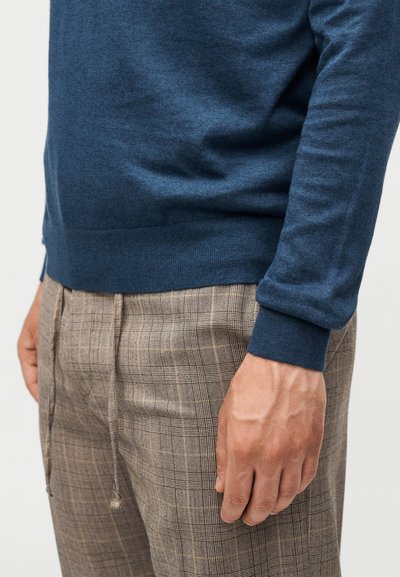 Άνδρας με μπλε πουλόβερ και καφέ καρό παντελόνι με κορδόνι, το δεξί χέρι χαλαρό στο πλάι, σε απλό φόντο.
