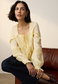 Żółta bluzka z długim rękawem, wykończona koronką, z wiązaniem z przodu i falbanowanymi mankietami, zestawiona z ciemnymi dżinsami i brązowymi butami.