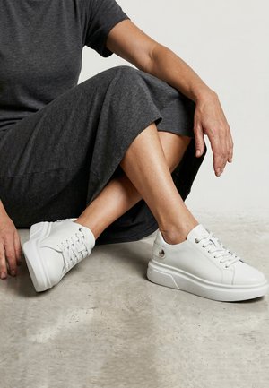 Osoba ubrana w ciemnoszara sukienkę, siedząca na podłodze ze skrzyżowanymi nogami, nosząca białe wiązane trampki na platformie.