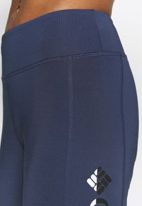 Námořnické modré sportovní legíny vyrobené z pružné tkaniny. Mají hladký, vysoký pas a bílé logo na dolní části nohy s geometrickými vzory.