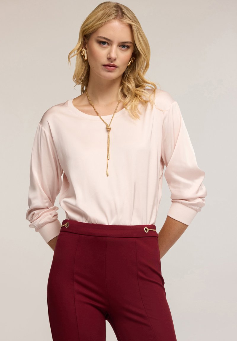 Blusa di raso rosa chiaro con maniche lunghe e scollatura rotonda, abbinata a pantaloni bordeaux su misura con dettagli in oro.