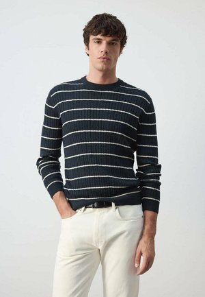 Hombre con un suéter azul oscuro con rayas horizontales blancas y pantalones claros, de pie frente a un fondo liso.