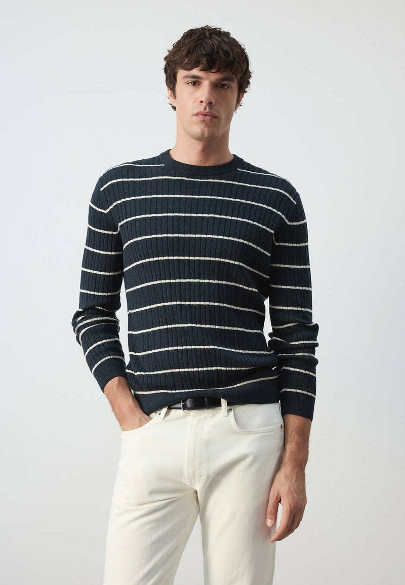 Homme portant un pull bleu foncé avec des rayures horizontales blanches et un pantalon clair, debout devant un fond uni.