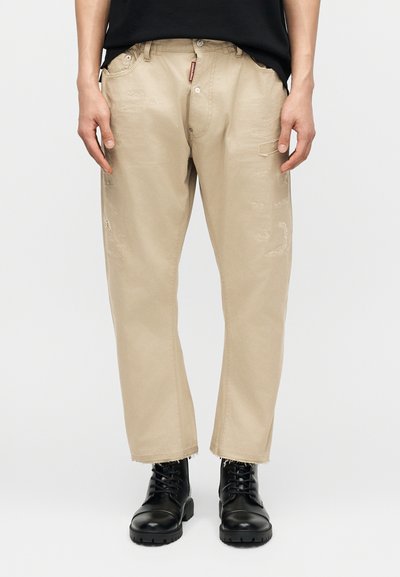 Pantalon beige coupe décontractée avec légère usure, associé à des bottines noires à lacets, porté par une personne debout devant un fond uni.