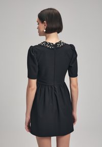 Krátké černé šaty s nabíranými rukávy, shromážděným pasem a dekorativním špičatým límcem. Hladká tkanina, délka po kolena, minimalistický design.
