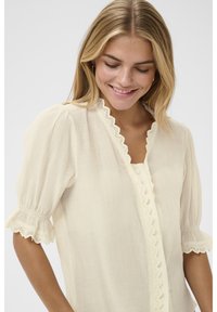 Ženska z blond lasmi, nasmejana, oblečena v kremno bluzo z valovitim čipkastim robom in rahlo nabranimi kratkimi rokavi, gleda navzdol.