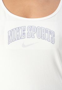 Bílý tílko s výšivkou "NIKE SPORTS" v purpurové barvě a malým stříbrným logem Nike. Kulatý výstřih a hladká bavlněná textura.