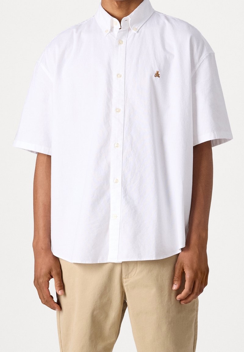 Chemise blanche à manches courtes avec boutons et une petite abeille brodée sur la poitrine gauche, portée avec un pantalon beige.