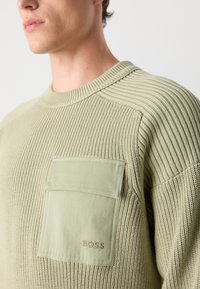 Pull en tricot texturé vert avec un col côtelé, une poche latérale et un logo "BOSS" brodé dans le coin de la poche.