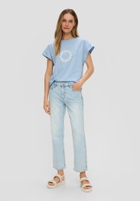 Lichtblauw katoenen t-shirt met korte opgerolde mouwen en een witte zonafbeelding. Draagt het met lichte spijkerbroek met rechte pijpen en beige sandalen.