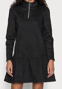 Černé džíny šaty s vysokým límcem a polovičním zipem, s dlouhými rukávy a rozšířeným volánovým lemem. Měkká textilie, ležérní design.