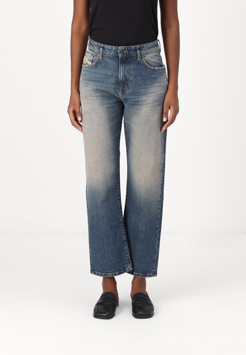 Personne portant un jean bleu délavé à jambes droites, un haut noir et des chaussures noires sans lacets, debout devant un fond blanc uni.