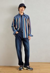 Πολύχρωμο ριγέ πουκάμισο με τζιν λεπτομέρειες, συνδυασμένο με σκούρα μπλε τζιν και πολύχρωμα αθλητικά παπούτσια, φορεμένο με σκούρο καπέλο.