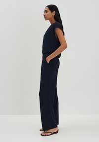 Marineblauwe jumpsuit met korte mouwen, ontspannen pasvorm, zijzakken en wijde benen, gecombineerd met zwarte platte sandalen tegen een effen achtergrond.