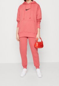 Kvinna i korallrosa Nike-hoodie och matchande byxor, som håller en liten glänsande röd handväska, med vita sneakers på en enfärgad bakgrund.