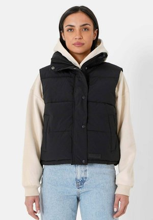 Veste matelassée noire avec col montant, boutons-pression et poches latérales, portée sur un sweat à capuche crème et un jean en denim bleu. Texture lisse.