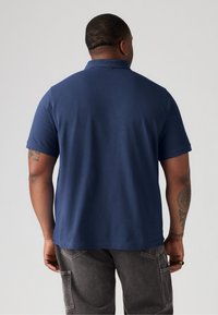Donkerblauw polo shirt met een kraag en korte mouwen, gemaakt van gladde stof. Heeft een relaxte pasvorm en een zichtbare tattoo op de arm.