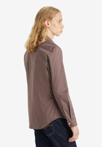 Brun langærmet skjorte med en glat tekstur, knaplukning ved håndleddene og en afrundet kant, båret med mørke jeans, set bagfra.