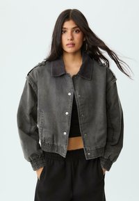 Grå denim bomberjakke med en kordfløjskrave, elastiske manchetter og kant. Har trykknapper og sidelommer for funktionalitet.