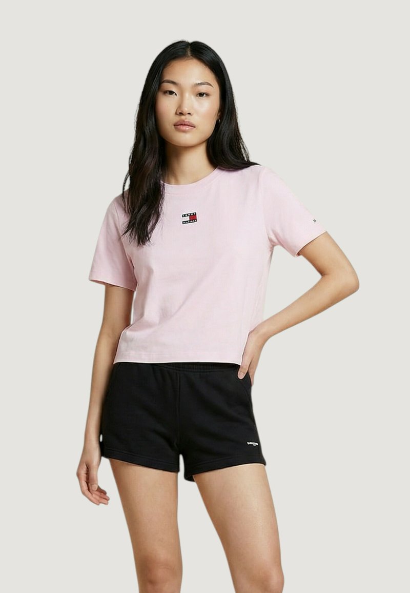 Woman with long dark hair wearing a pastel pink cropped T-shirt and black athletic shorts, standing with one hand on her hip.