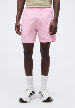 Mann trägt hellrosa Shorts, weiße Sportschuhe mit schwarzen Akzenten, kombiniert mit weißen Kniestrümpfen, steht vor einem schlichten Hintergrund.